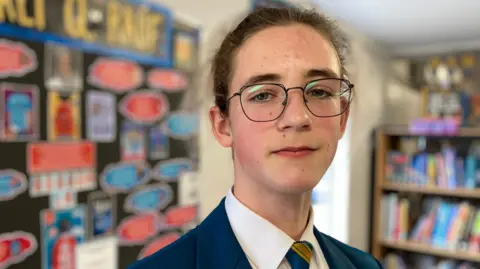 Roman is looking directly at the camera. He has dark hair and is wearing glasses, a blue blazer, white shirt and tie with blue and yellow stripes. 