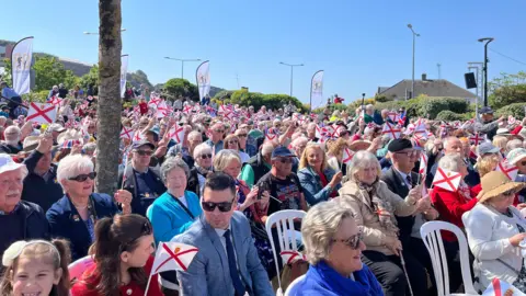 A crowd of people sat on white chairs waiting to hear speeches from island officials in Jersey on Liberation Day in 2025