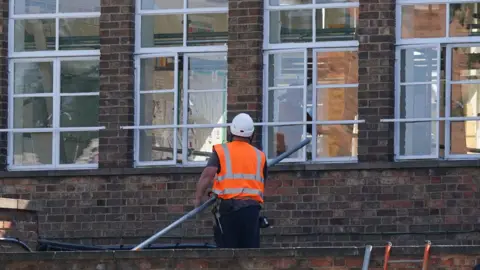A construction worker in a hard hat and orange safety vest stands outside the brick building of a school, holding a long pole while working on or inspecting the exterior window frames. Several large windows reflect the surroundings, and scaffolding or support bars are positioned across them.