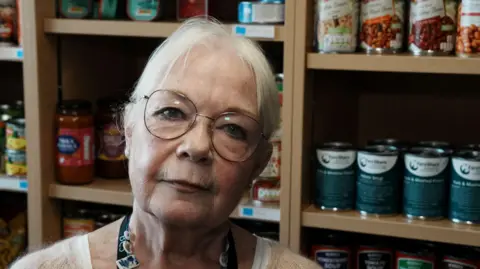John Fairhall/BBC A head and shoulders image of a woman wearing glasses and a white top. She is looking into the camera while standing in front of shelving on which there are tins of cans goods.