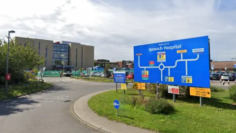 A general view of the entrance of Ipswich Hospital. It shows a road sign directing drivers around the hospital site. A hospital building can be seen in the background as well as a car park. 