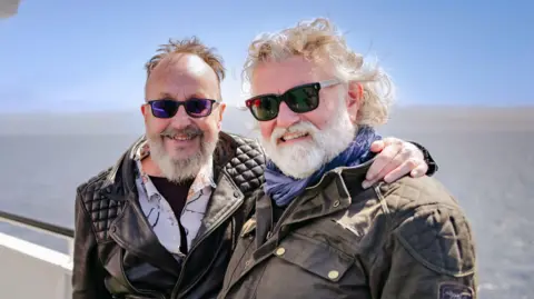 Hairy Bikers Dave Myers and Si King are standing on a boat out in the water and smiling at the camera. Myers, on the left, has his arm around King and is wearing a black leather jacket. King is wearing a brown jacket and they are both wearing sunglasses.