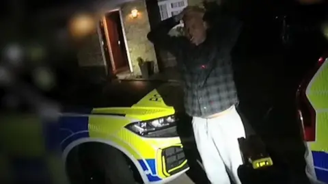 Jeremy Heath has his hands on his head, wearing a dark-coloured check shirt, and standing outside a house in darkness. There are two marked police cars parked either side of him. He appears to have a cigarette in his mouth.