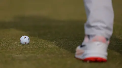 Getty Images Close up of a white golf ball with four distinctive triangle pen markings. A blurred white golf shoe, with orange soles, is blurred in the foreground 
