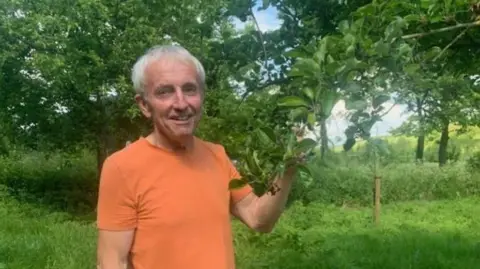Oliver Dowding Oliver standing in a mid-shot beside a tree in an orchard - he is holding onto the branch of an apple tree, and is wearing an orange T-shirt. He has white hair. 