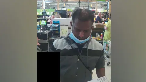 CPS Mathiyaparanam is pictured at a supermarket self-service checkout buying a box of knives. He has short black hair and is wearing a grey striped zipped-up top, black t-shirt and a blue face mask.