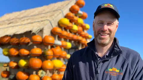 Farmer Jason Butler stands by a wooden, house-shaped structure, which is covered in orange and yellow pumpkins, against a backdrop of a deep blue sky.  He is wearing a blue cap and fleece, both featuring a pumpkin logo and the words "The Pumpkin Patch at Bewholme".