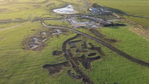 Holly Wilkinson An aerial view of a flat landscape with a large pond and other rivulets nearby. Some agricultural buildings can be seen on the upper right of the image.