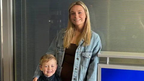 A young boy who has dark blonde hair is standing with him mum who has long blonde hair, a black top and blue denim jacket. She has her arm around him