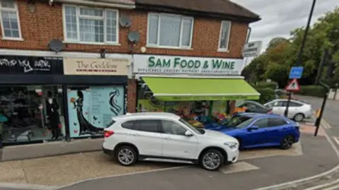 Google A parade of shops on a corner includes Sam Food and Wine. There is a driveway with cars immediately in front of the shops, then a kerb and a strip of pavement and then another kerb and the main road.