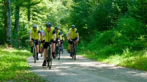 Cyclists on a country track surrounded by green trees and patches of sunlight