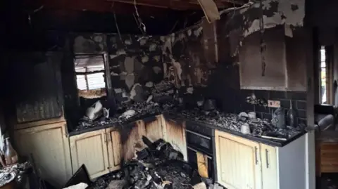 A square-shaped kitchen layout with white cupboards badly burned, charred and damaged by by fire.