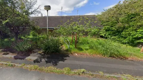 A red brick building in the background that is surrounded by trees and bushes. The pavement runs along the bottom of the image. There is a lampost in the middle as well as metal railings that run either side of a path leading to the building. 