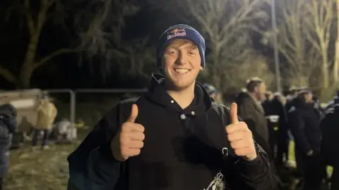 Jasmine DuFraisse/BBC Angry Ginge, wearing a dark blue woolly hat with the Red Bull logo and a black hoodie, smiles at the camera while giving a thumbs-up gesture with both hands.