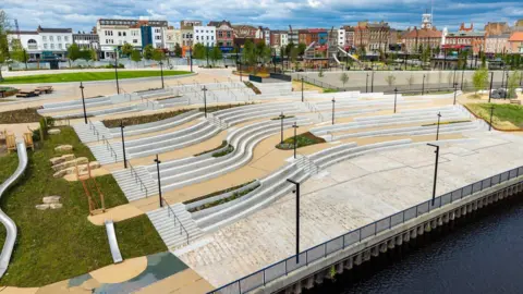 An aerial view of the white steps of the parks amphitheatre which faces on to the river. There are winding paths, slides and spots of greenery dotted around the development. High Street in the background is lined with multi-storey brick buildings in different colours.