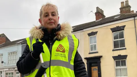 A woman wearing a fluorescent yellow bib which includes a "poo emoji logo - a triangular brown poo shape with eyes and a mouth - and the words 'Town S.M.A.R.T. Dog Poo Patrol'. She has blonde hair, tied back, and is wearing a black parka-style coat beneath the vest. She is holding the handle of a litter-picking device and standing in a street with a terrace of colourful properties behind her.