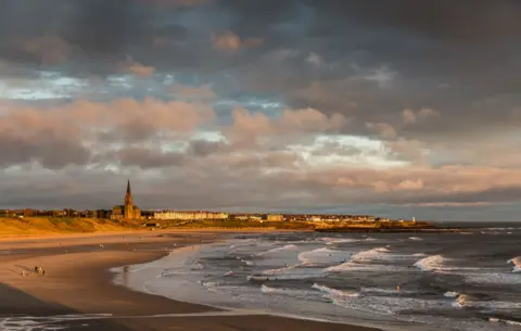 Getty Images Longsands beach. It is a great sweep of golden sand with waves breaking along it, and buildings on the cliff beyond including a long terrace of large homes and a church with a spire bathed in sunlight.