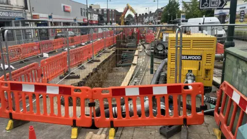 A large trench has been dug out of the road with construction items like diggers visible next to it. There is metal fencing around the hole and orange barriers.