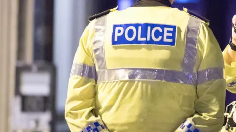 The back of a male police officer, who is wearing a yellow long-sleeved hi-vis jacket with reflective silver stripes and a blue badge that reads "POLICE".