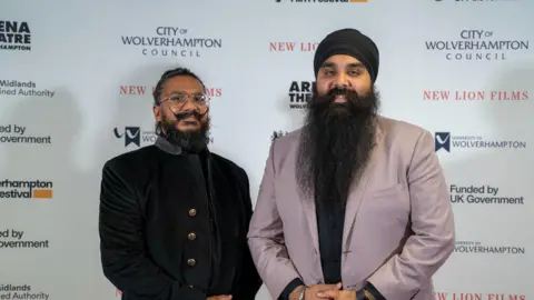 Arun Kapur and Gurjant Singh - Two men, one in a black outfit, glasses handlebar moustache and the other wearings a pink blazer, a black turban and has a long black beard. They stand against a white wall with logos