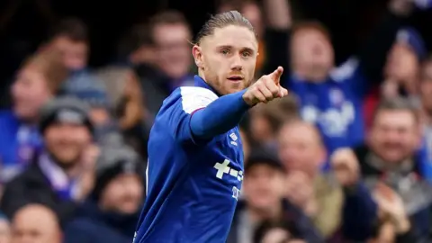 PA Media A football player celebrating after a goal. He is pointing his finger out as he runs. He is wearing a blue football shirt, and he has long brown hair that has been tied back.