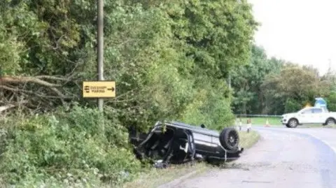 West Mercia Police The upturned remains of the BMW are at the side of the road, with its rear lodged into trees. A lamp-post is just in front of the car, and in the distance a white pick-up truck can be seen on a roundabout where traffic cones can be seen