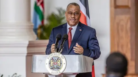 Haiti's former prime minister Garry Conille standing behind a lectern with two microphones attached to it. His hands are in front of him, and there are two out of focus flags in the background. The lectern says Republic of Kenya on the front of it. 