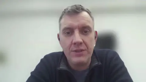 Nerijus Gecas was interviewed via Zoom. His background is obscured but is a largely white room. He is a white man with short greying hair, and is wearing a black, zip-up fleece and can be seen to have black in-ear headphones on. 