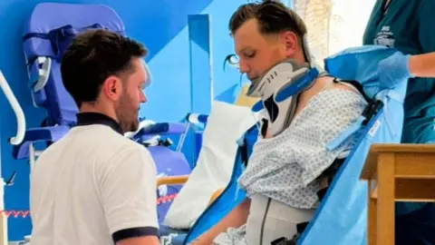Spinal Research Sam Baldwin in a neck-brace and hospital scrubs. He is sat on a chair with a physiotherapist moving his legs.