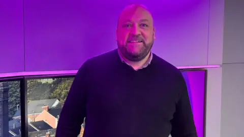 A man with a shaved head, and short beard, wearing a dark blue jumper over a shirt. He is smiling at the camera