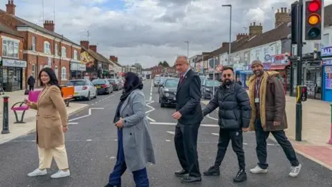 Peterborough City Council Shabina Qayyum in a beige coat and white trousers, Noreen Bi in a grey coat, black headscarf and blue trousers, Angus Ellis is a grey suit, Asim Mahmood in a black puffer jacket and black jeans and Zameer Ali in a brown coat, crossing the road, while looking into the camera.