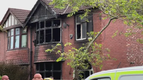 The house in Chorlton, its front windows are broken and black with the smoke from the fire 