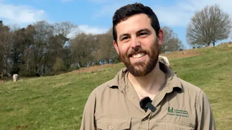 Rob Lee wearing a brown shirt, standing in Beacon Hill Country Park, in Leicestershire, with trees in the background on a sunny day.