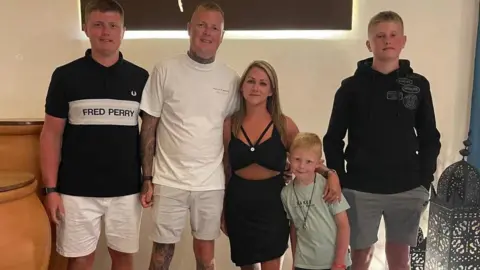 Family handout via West Yorkshire Police Koby Jones (far left) pictured with his family. He is wearing white shorts and a T-shirt printed with the words Fred Perry. He is stood next to a man wearing a white T-shirt and light shorts. Also stood in a line is a woman, wearing a black dress with her arm around a young child. There is a teenage boy stood at the end of the line wearing grey shorts and a black hoodie.