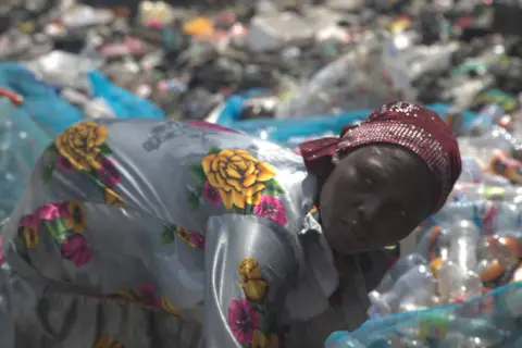 Abiba Alhassan, seorang wanita yang mengenakan gaun bermotif bunga dan jilbab, sedang memilah botol plastik bekas di samping tempat pembuangan sampah Agbogbloshie di Accra