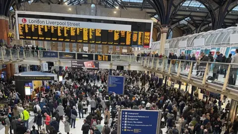 James Harrod An overheard shot of a very busy concourse at London Liverpool Station, under a glazed roof. There are crowds of people in the foreground and stretching back towards the large electronic train information sign, which has bright yellow lettering on it, with London Liverpool Street written across its top. 
