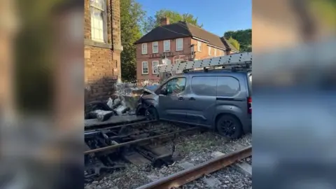 Network Rail A dark grey van with ladders on the roof which has crashed into a level crossing. The bonnet of the van is smashed.