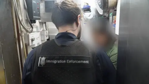 The back head and shoulders of an immigration officer, with a black gilet showing the badge 'immigration enforcement on the back. In the background there is a blurred out photo of someone wearing a dark green top. They are facing each other in a small cupboard area with switches and wires on the left side.