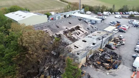 A drone shot from above a damaged unit. There are charred buildings and vehicles on the right, and big holes in the roof.