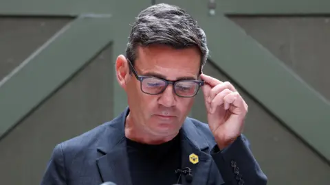 Reuters Andy Burnham looks down, holding the side of his glasses. He wears a blue jacket, blue top and a yellow badge with a bee symbol on it.