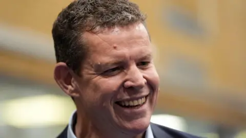 The image is a close-up of Plaid Cymru's leader Rhun ap Iorwerth. He is looking away from the camera and is smiling. 