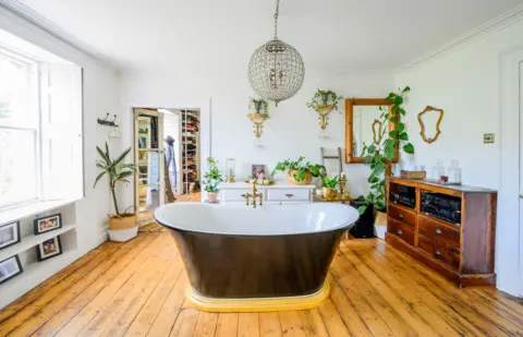 IWC Media A bright, spacious bathroom features a freestanding black bathtub placed centrally on warm wooden floorboards.
White walls are decorated with mirrors, plants, and shelves, while a large window lets in abundant natural light.
A chandelier hangs above, and vintage furniture and accessories add a stylish, eclectic touch to the room.