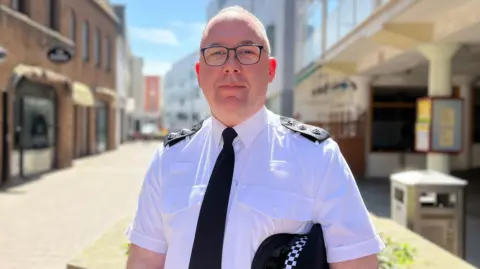 David is wearing a white short‑sleeved uniform shirt with shoulder epaulettes and a black tie stands outdoors on a pedestrian street, holding a black hat with a checkered band. Buildings line both sides of the street, and the background is softly blurred under bright daylight.