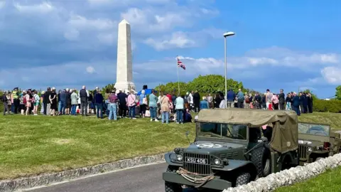 Portland Town Council A large group of people gathered around a war memorial on an area of grass. In the foreground is a road and two parked world war two-era army vehicles. It's a sunny day and the sky is blue with scattered clouds.