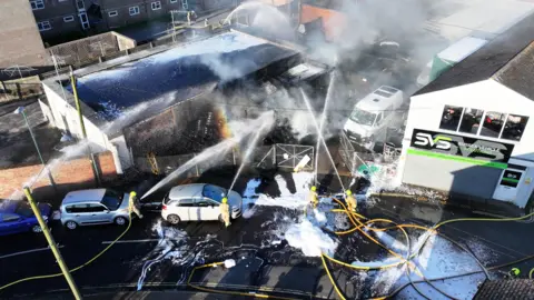 Four firefighters are seen from above spraying water on a smoldering industrial building. 