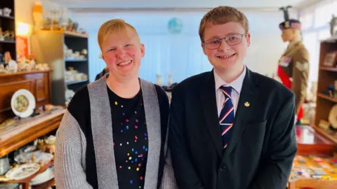 Woman, smiling, wearing grey and black cardigan, over a black top with bright dots, and boy, 16, smiling, wearing glasses, black jacket, white shirt, red white and blue striped tie, holding medals, with antiques in the background.