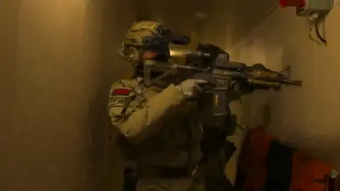 A screengrab from a video which depicts an armed forces personnel in tactical gear holding a gun. They are in a small corridor and wearing a helmet and gloves with most of their face covered.