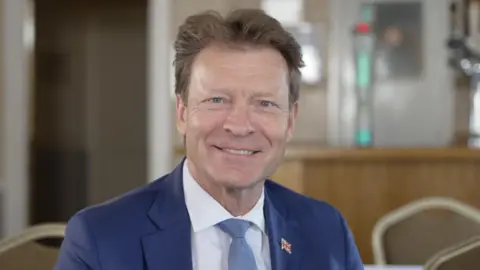 A man smiles as he sits in a bar room, the background blurred. He has brown hair, swept back and wears a blue suit over a white shirt and blue tie. He also wears a Union flag lapel badge.
