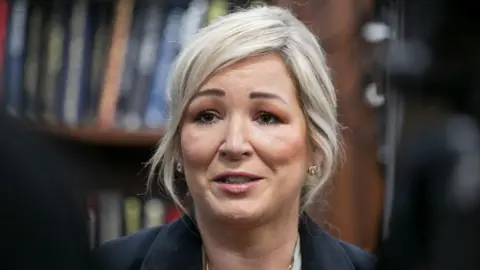 Michelle O'Neill, who has blonde hair, wears a dark jacket and gold earrings as she speaks to the media.