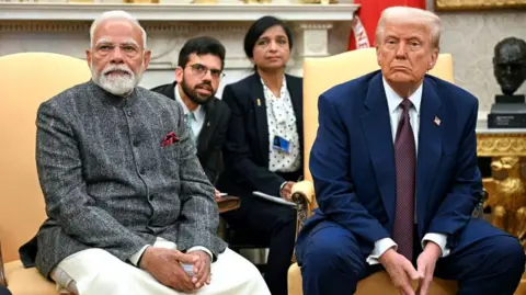 Getty Images US President Donald Trump speaks with the press as he meets with Indian Prime Minister Narendra Modi in the Oval Office of the White House in Washington, DC. Both look at the camera with folded hands. Modi is wearing a traditional Indian kurta with a jacket, while Trump is in a navy blue suit and tie.